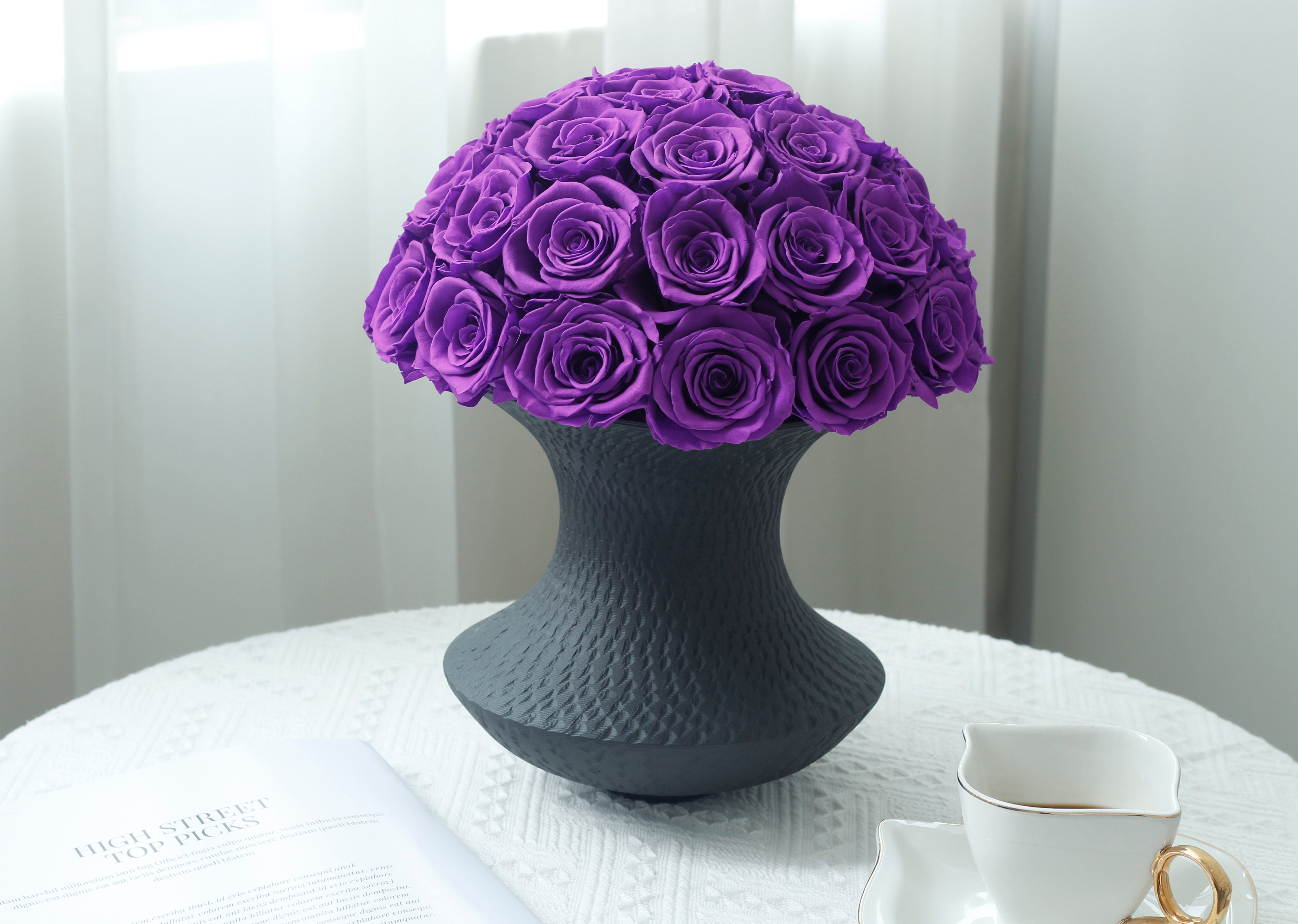 Elegant arrangement of preserved purple roses in a textured black vase, placed on a white table with a coffee cup and a magazine nearby